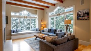 A cozy living room with a sofa, a coffee table, and a lamp. There are large windows with a view of snow-covered trees. - Ski-In/Ski-Out | Modern 4BR | Private Hot Tub | Double Garage | Managed by Aloha Whistler (Whistler)