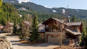 A two-story lodge with a garage, surrounded by a forest and mountains in the background. - Ski-In/Ski-Out | Modern 4BR | Private Hot Tub | Double Garage | Managed by Aloha Whistler (Whistler)