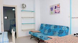 A room with a blue sofa, a bed with a blue and white checkered bedspread, a small table with a checkered tablecloth, and a wall-mounted fan. Shelves are attached to the wall above the sofa. - One bedroom apartment with WiFi (CONIL DE LA FRONTERA)