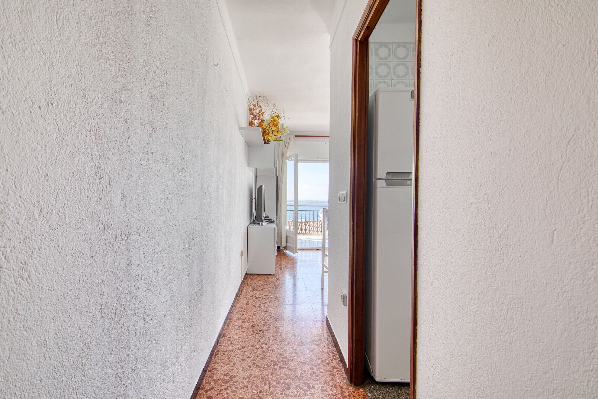 A hallway with a white textured wall, a tiled floor, and a door leading to a balcony with a view.