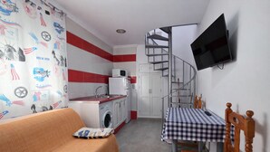 Un espacio habitable compacto con un sofá, una mesa con mantel a cuadros, una lavadora, un microondas, un televisor montado en la pared y una escalera de caracol.