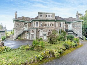 Un edificio de piedra con una entrada central, una escalera que conduce a un balcón y un jardín circundante.