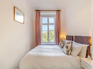 A bedroom with a large bed, a window with curtains, a bedside table with a lamp, and a framed picture on the wall.