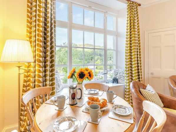 Un comedor con una mesa lista para el desayuno, una ventana con vista a la vegetación y cortinas estampadas.
