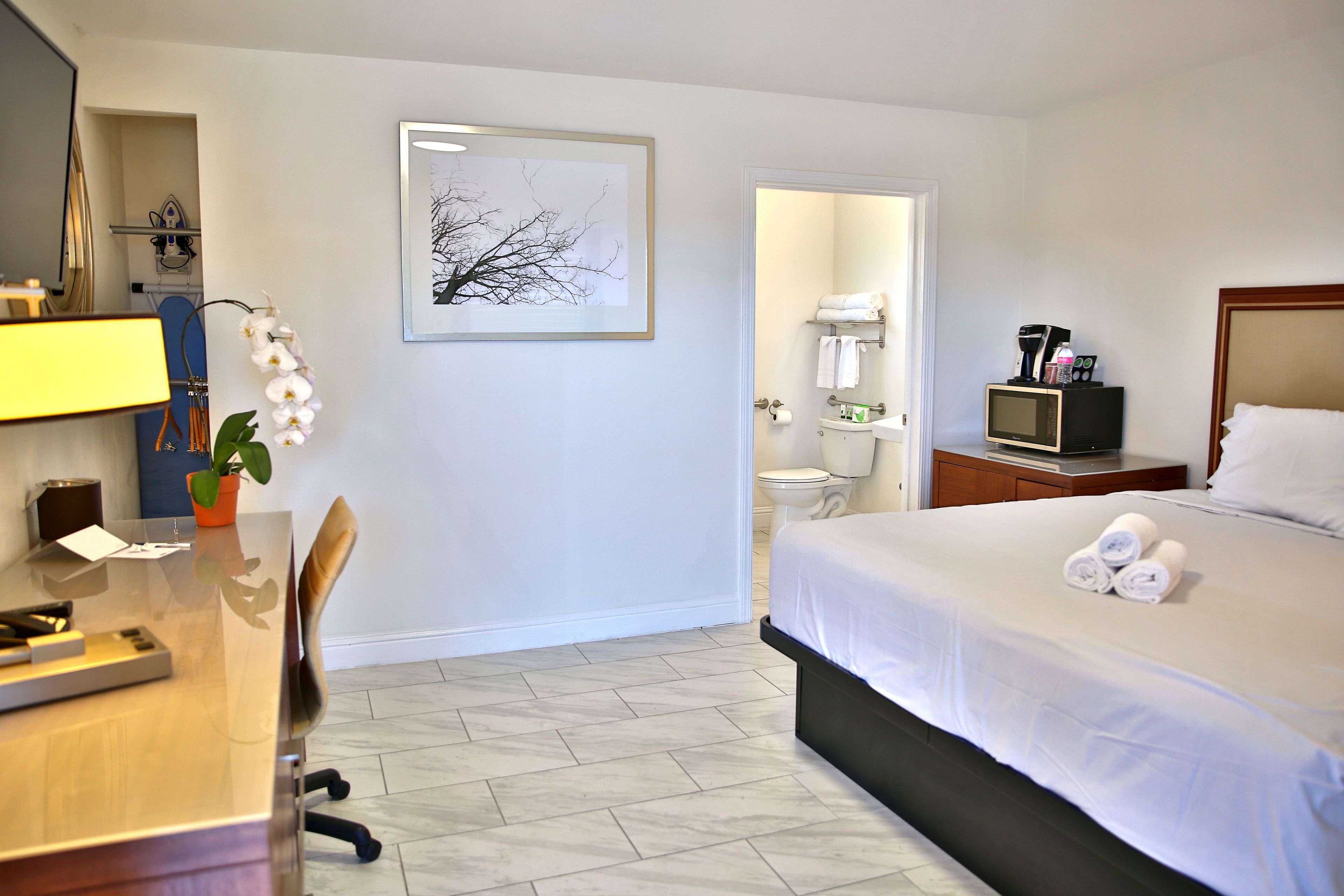 Habitación de hotel con cama, escritorio con computadora, silla, televisor, cafetera y un cuadro en la pared.