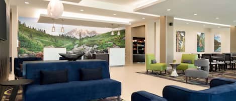 A modern hotel lobby with a blue sofa, green armchairs, a dining area, and a mural of a mountain landscape.