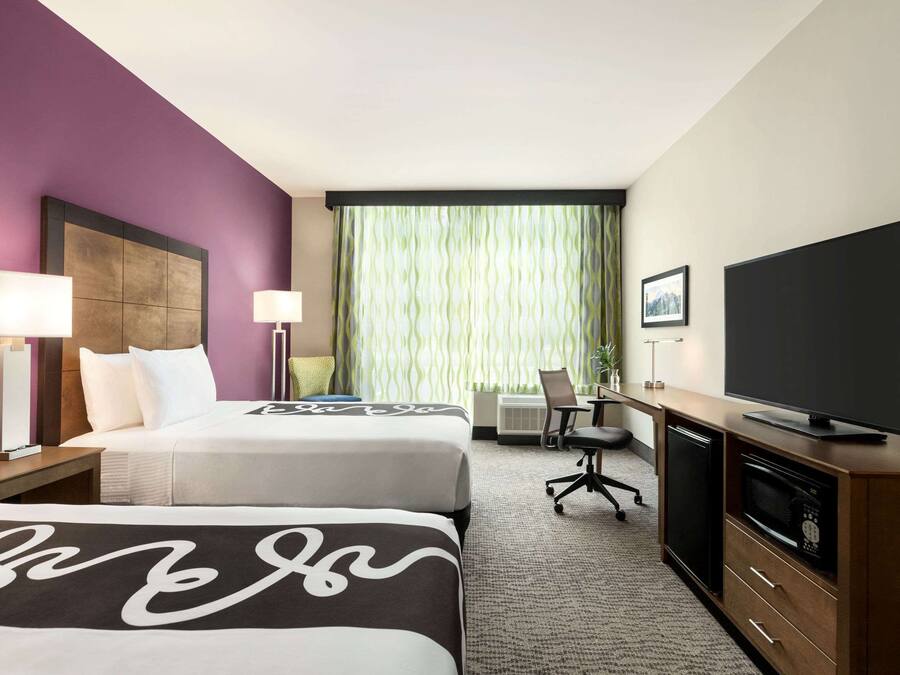 A hotel room with two beds, a desk, a television, and a window with curtains.