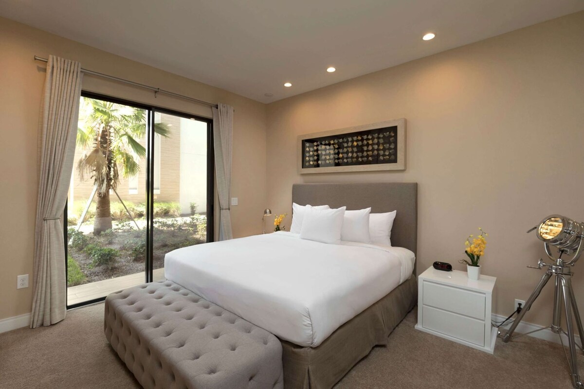 A hotel room with a large bed, a bedside table with a lamp, a small table with flowers, and a view of palm trees through the window.