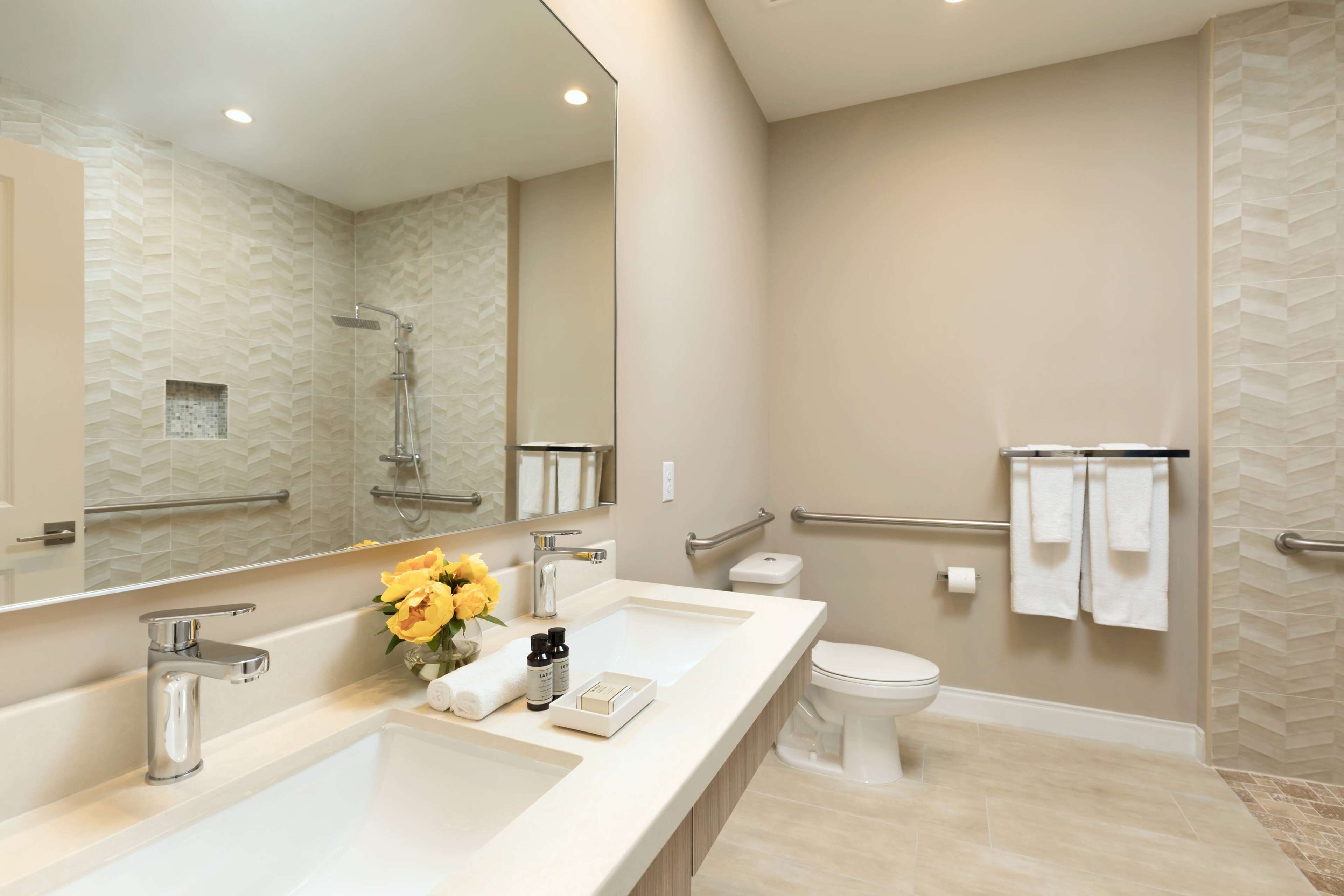 A modern bathroom with a large mirror, two sinks, a shower, and a toilet.