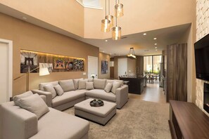 Una sala moderna con un amplio sofá sectional en forma de L, una mesa de centro y un televisor sobre un mueble de madera. Se observa una cocina al fondo con una mesa de comedor y sillas.