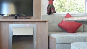 Et moderne interiør med et fladskærms-tv monteret over en indbygget pejs, en sofa med en rød pude, og et vindue med udsigt til grøn vegetation.