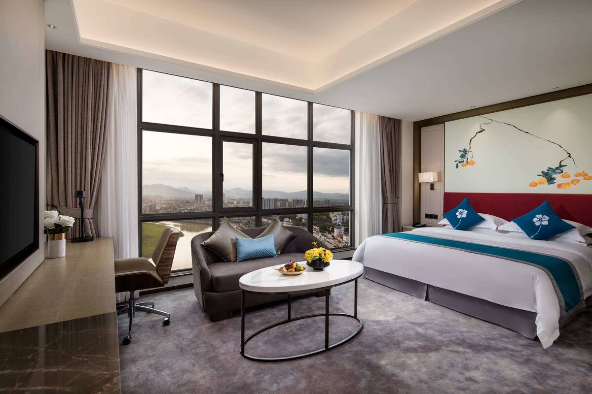 Een hotelkamer met een groot raam, een bed, een bank, een salontafel en een bureau.