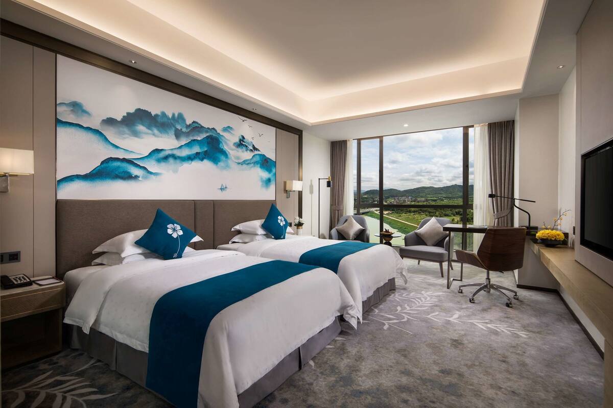 Een hotelkamer met twee bedden, een groot schilderij aan de muur, een bureau met een lamp en uitzicht op het landschap door het raam.