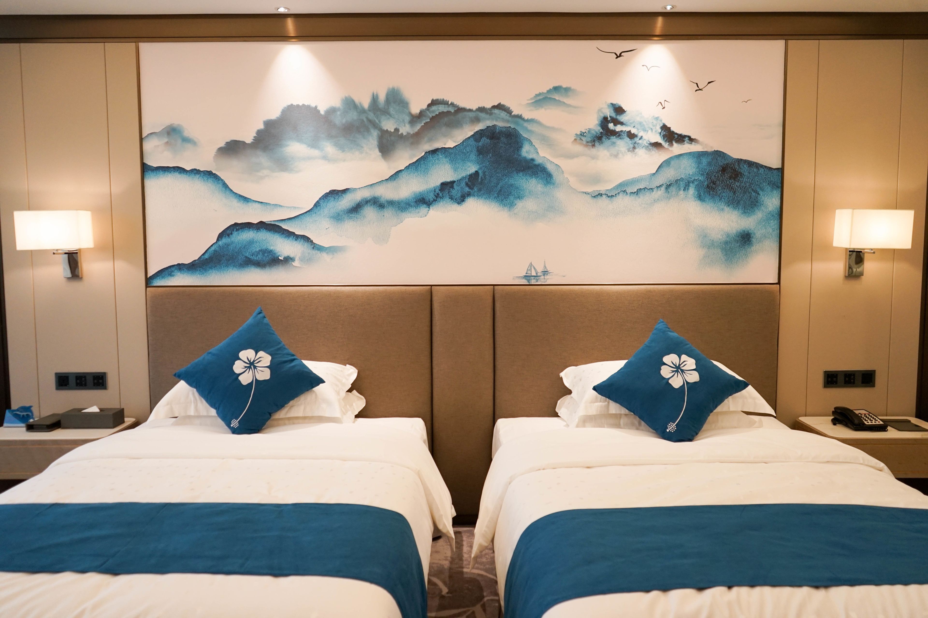 A hotel room with two beds, each with a blue floral pillow, a large wall painting of mountains, and two bedside lamps.