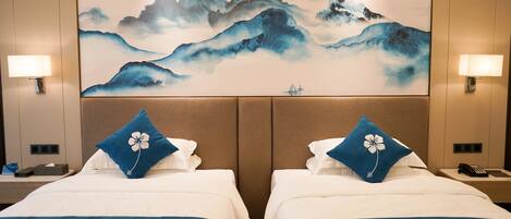 A hotel room with two beds, each with a blue floral pillow, a large wall painting of mountains, and two bedside lamps.