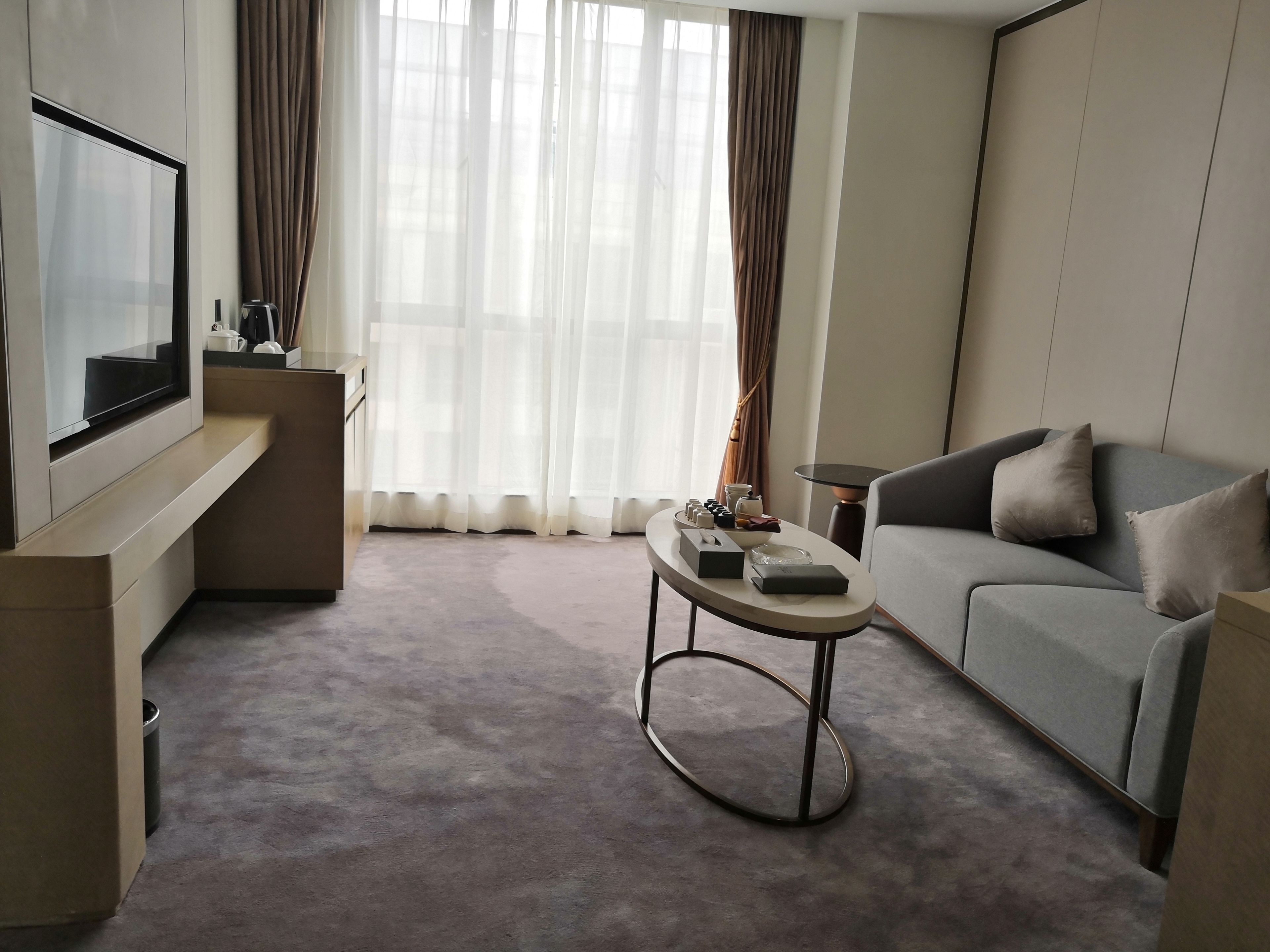 A modern hotel room with a flat-screen TV, a grey sofa, a coffee table, and a large window with sheer curtains.