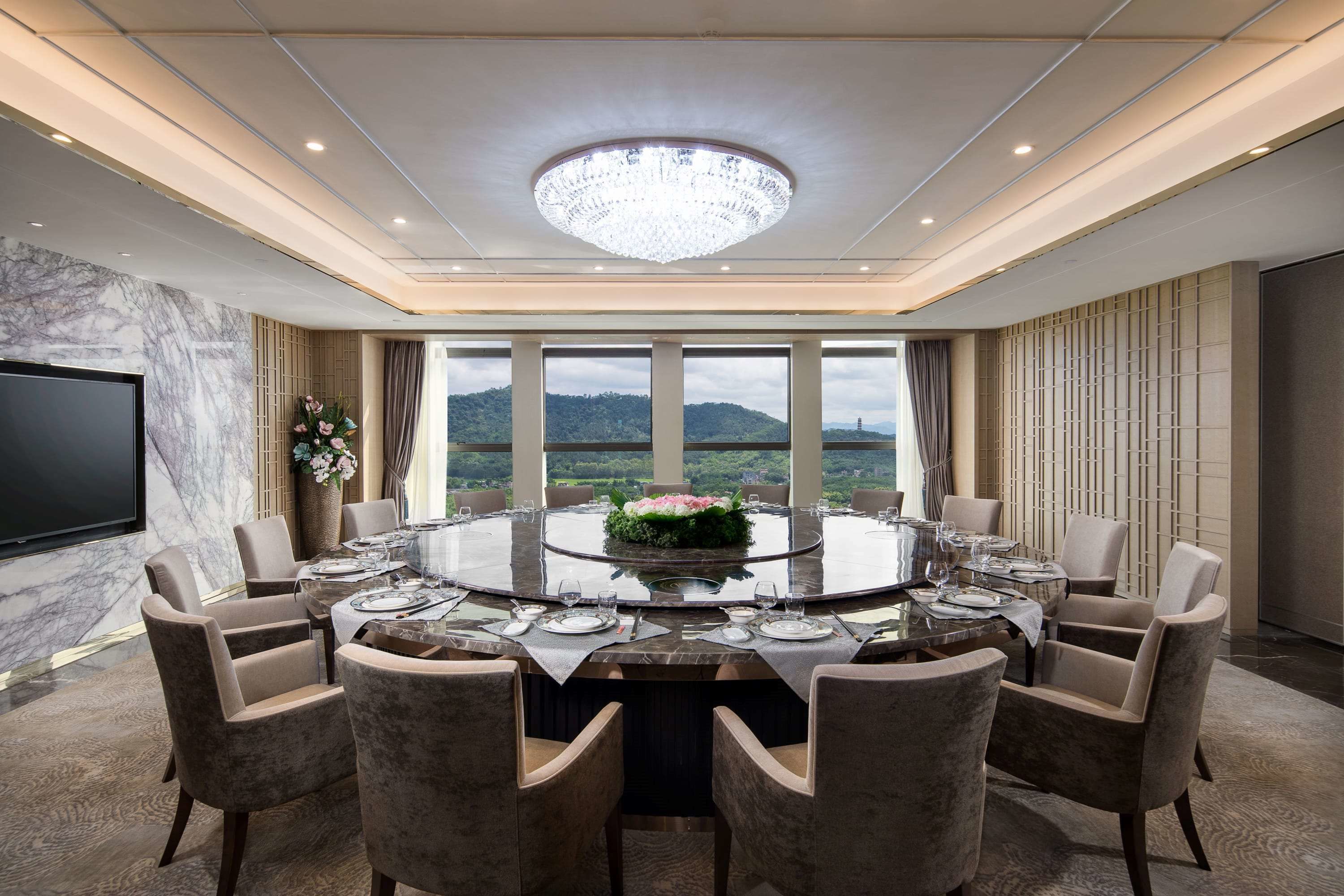 A conference room with a round table, chairs, a chandelier, a flat-screen TV, and a view of the outdoors.