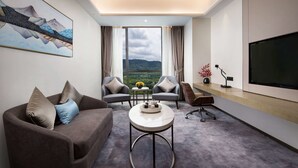 Ein modernes Hotelzimmer mit einer Couch, zwei Sesseln, einem Couchtisch, einem Fernseher und einem großen Fenster mit Blick auf die Berge.