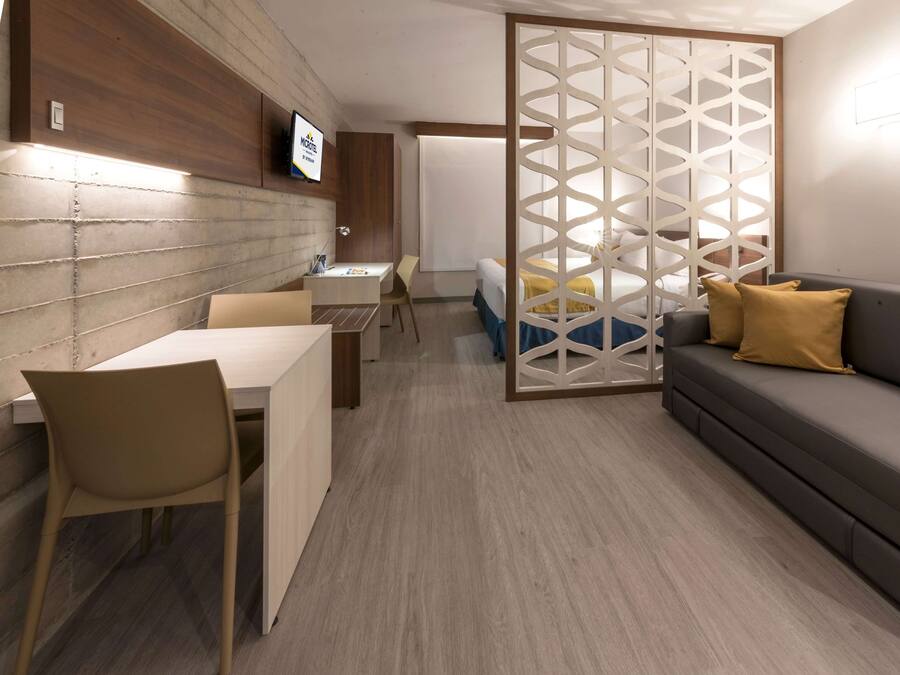 A modern hotel room with a wooden accent wall, a flat-screen TV, a sofa with yellow pillows, a desk with a chair, and a bed with a blue and yellow bedspread.