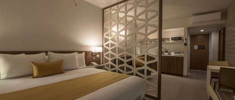 Habitación de hotel con una cama grande, cabecera de madera, mesita de noche con lámpara, separador decorativo, kitchenette con microondas y un pequeño comedor.