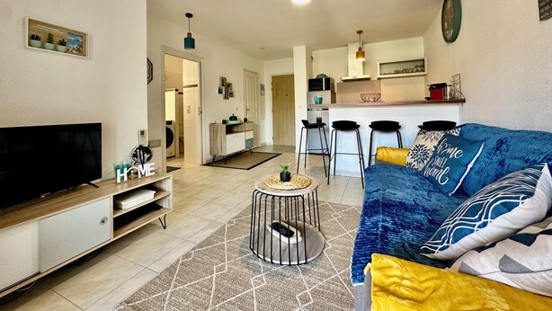 Uma sala de estar moderna com televisão, sofá e mesa de centro. Cozinha visível ao fundo, com banquetas de bar.