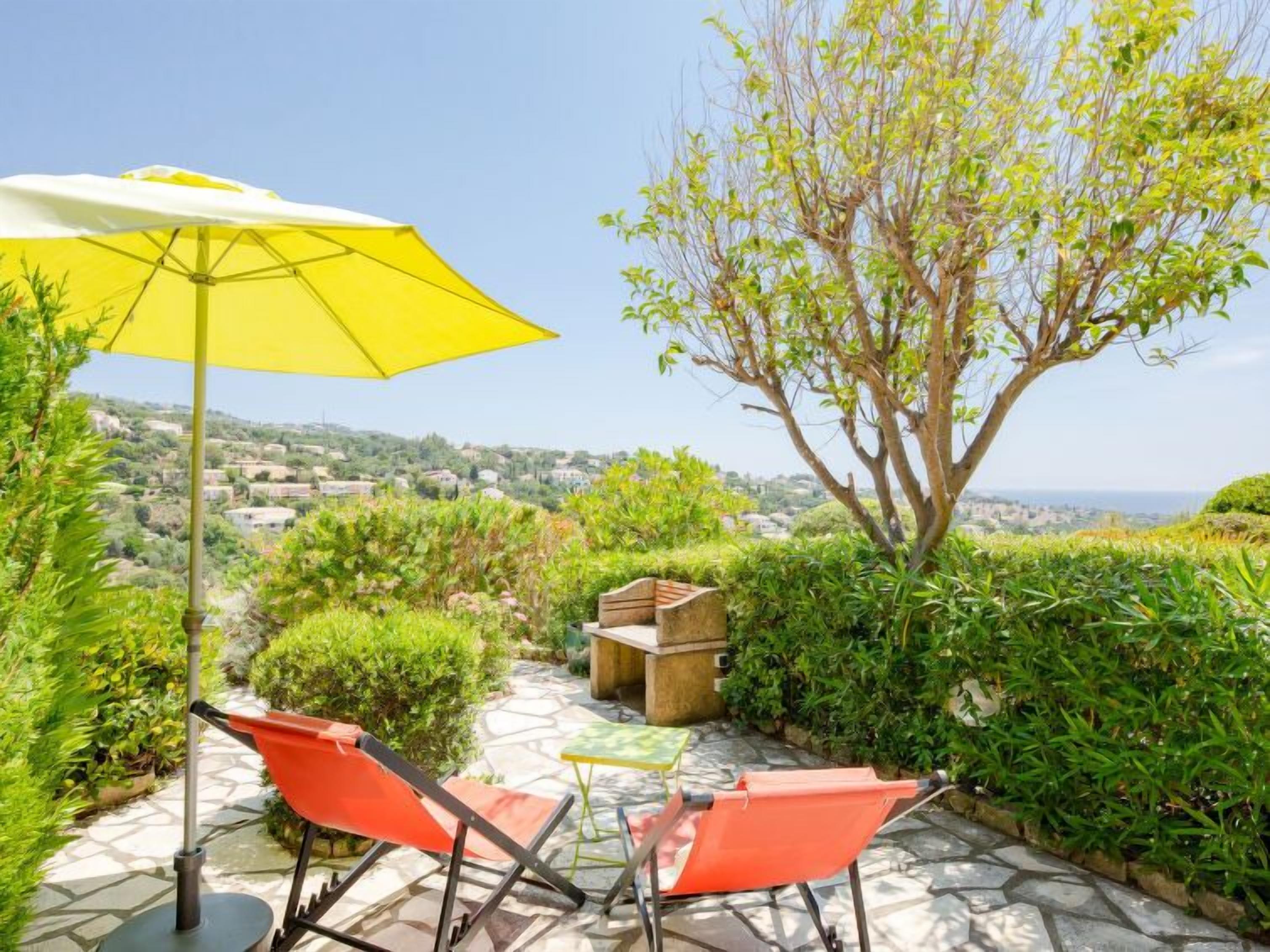 A patio with a yellow umbrella, two red chairs, and a view of a town and sea.