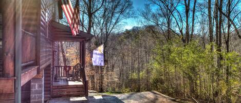 A wooden cabin with a porch, two flags, and a forested backdrop.