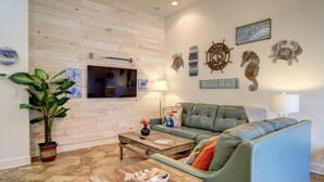 Ein Wohnzimmer mit einer Holzvertäfelung, einem grünen Sofa, einem Couchtisch, einem Fernseher und Wanddekorationen, darunter ein Schild mit der Aufschrift 'PRIVATE BEACH'.