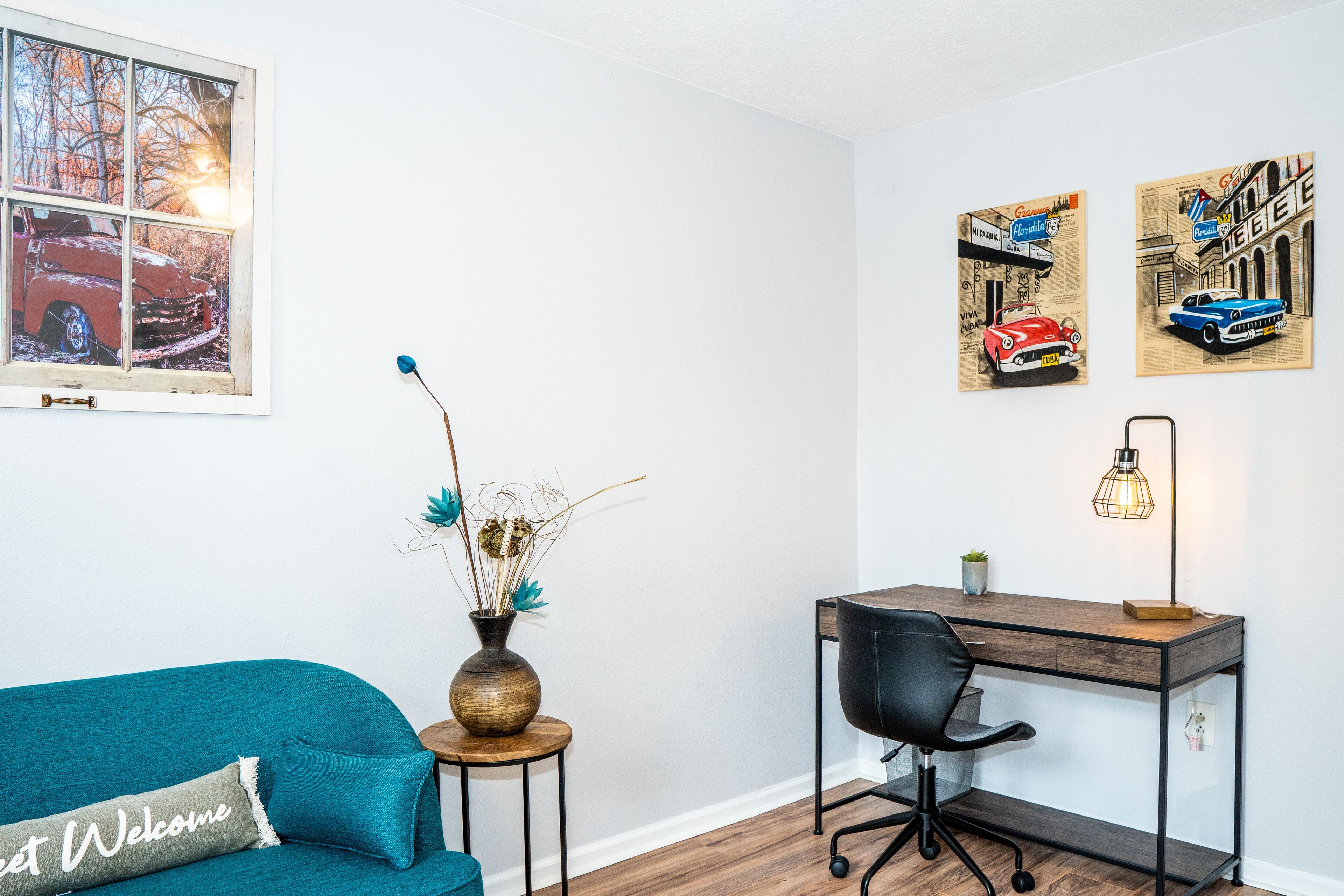 Ein Zimmer mit einem türkisfarbenen Sofa, einem Holztisch, einem schwarzen Bürostuhl, einer Vase mit Trockenpflanzen und zwei gerahmten Automobil-Postern an der Wand.
