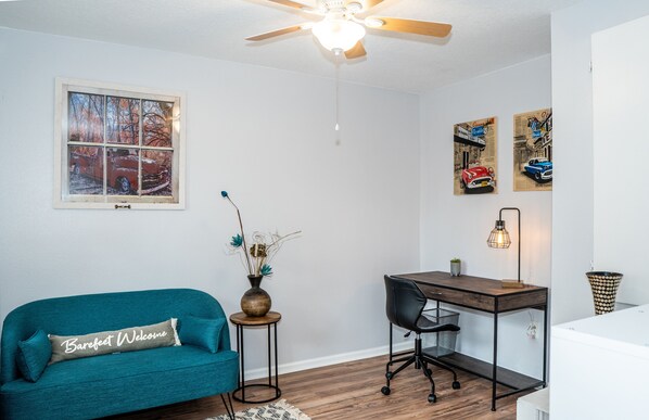 A room with a teal sofa, a wooden desk, a ceiling fan, and wall art.