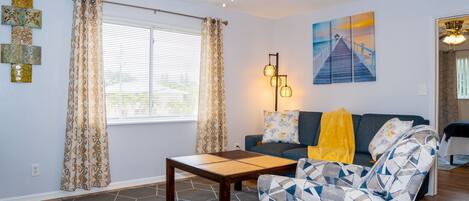 Una sala de estar con un sofá, un sillón estampado, una mesa de centro y un cuadro en la pared.
