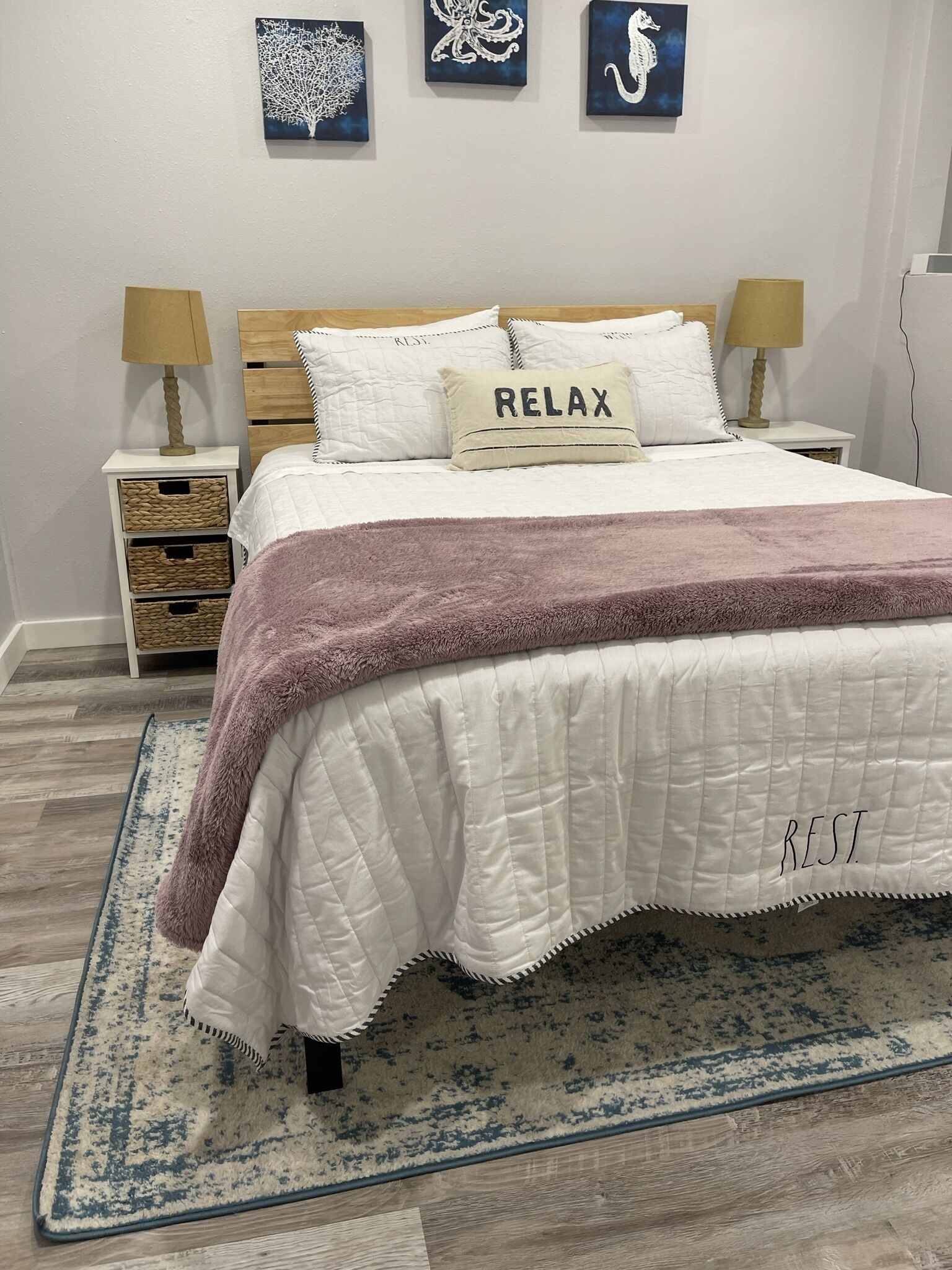 A bedroom with a bed, a wooden headboard, two bedside tables with lamps, a wall with three artworks, and a patterned rug.