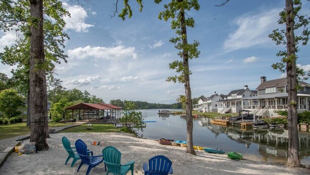 A lakeside view with a sandy beach, blue Adirondack chairs, a covered pavilion, and a dock with boats.