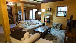 A cozy living room with a sofa, armchair, coffee table, and a television. There is a wooden bookshelf and a window with a view. - Cozy mountain log chalet overlooking the slopes of Hatley Pointe Ski Resort.  (Mars Hill)