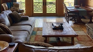 A cozy cabin interior with a wooden ceiling, a leather sofa, a dining table, and a large window overlooking greenery. - Cozy mountain log chalet overlooking the slopes of Hatley Pointe Ski Resort.  (Mars Hill)