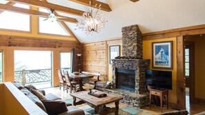 A cozy lodge interior with a stone fireplace, wooden furniture, and a large window overlooking a balcony. - Cozy mountain log chalet overlooking the slopes of Hatley Pointe Ski Resort.  (Mars Hill)
