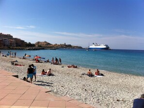 Ein Strand mit badenden und sonnenden Personen, im Hintergrund ein großes Kreuzfahrtschiff und Gebäude entlang der Küste.