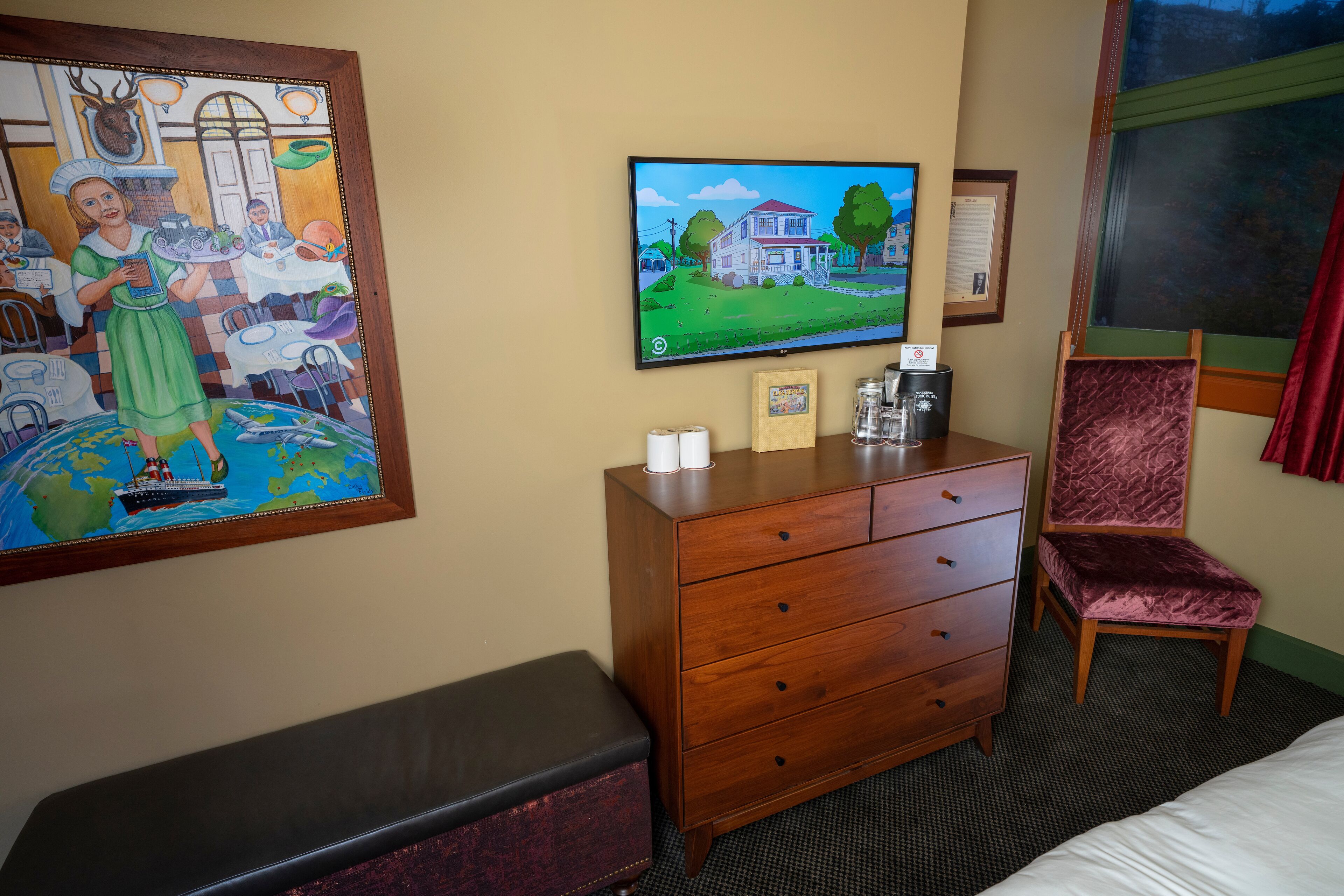 A hotel room with a wooden dresser, a television mounted on the wall, a framed painting, a red velvet chair, and a bed.