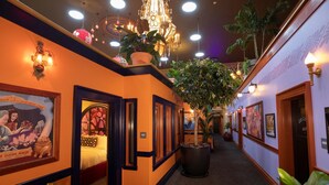 A hallway with a warm orange wall, black trim, and a large potted plant. There are framed pictures on the walls and a chandelier hanging from the ceiling. - McMenamins Elk Temple (Tacoma)