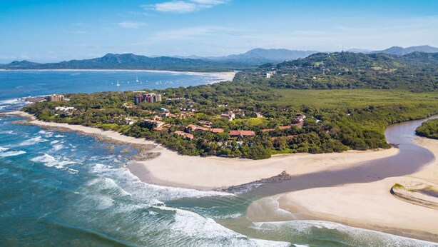 Une vue aérienne d’une ville côtière, avec une plage, des maisons et des montagnes en arrière-plan.