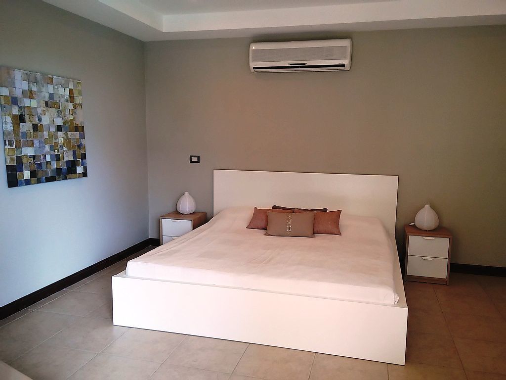 A modern bedroom with a large bed, a wall-mounted air conditioner, a decorative mosaic on the wall, and a nightstand with a lamp.