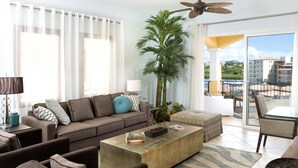 A living room with a sofa, coffee table, and chairs, a large plant, and a view of the city through large windows. - Agua Azul by Island Properties Online (Lowlands)
