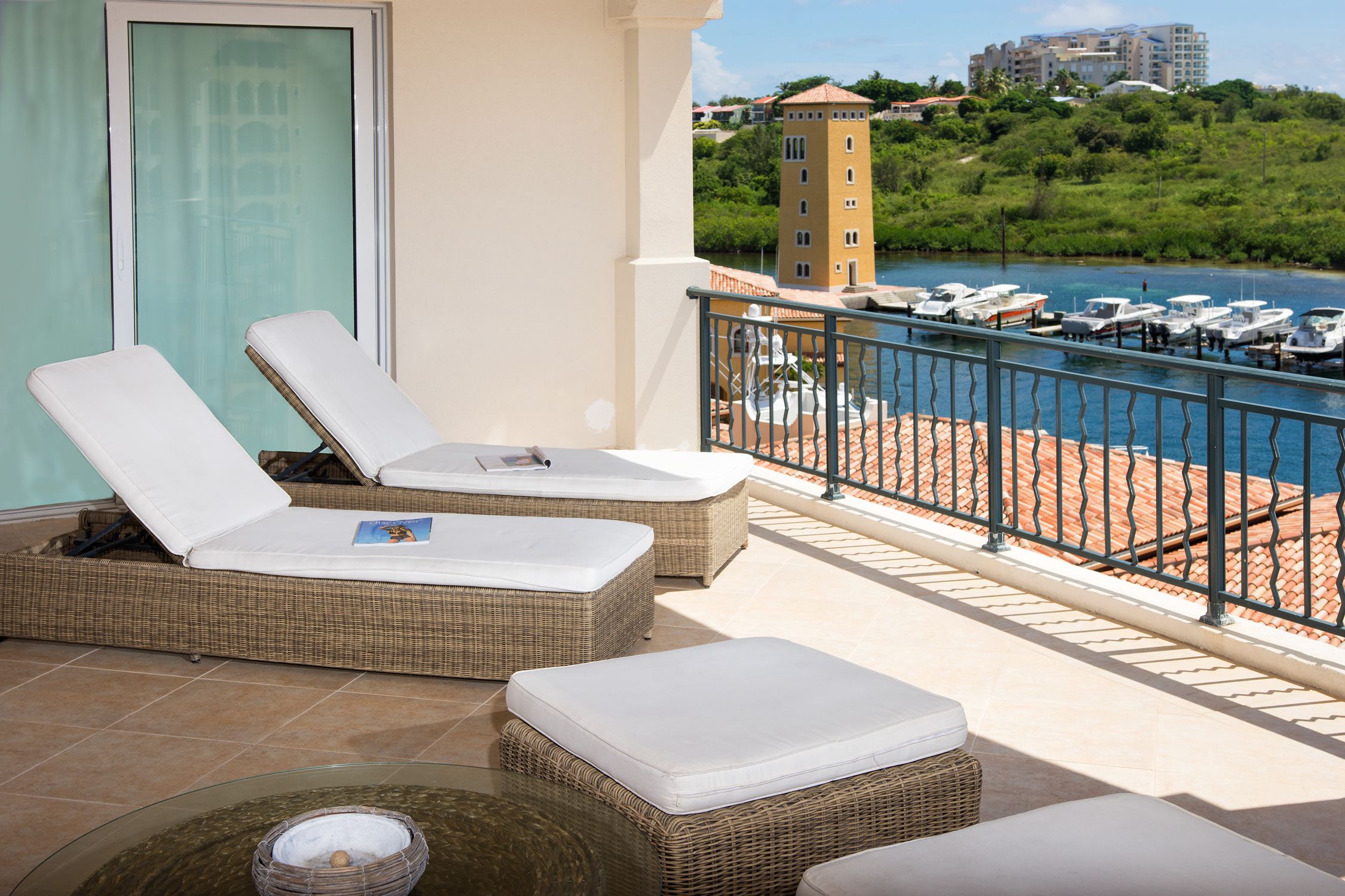 Un balcon avec deux fauteuils de jardin en osier et une petite table, donnant sur une marina où sont amarrés des bateaux, avec un bâtiment visible au loin.