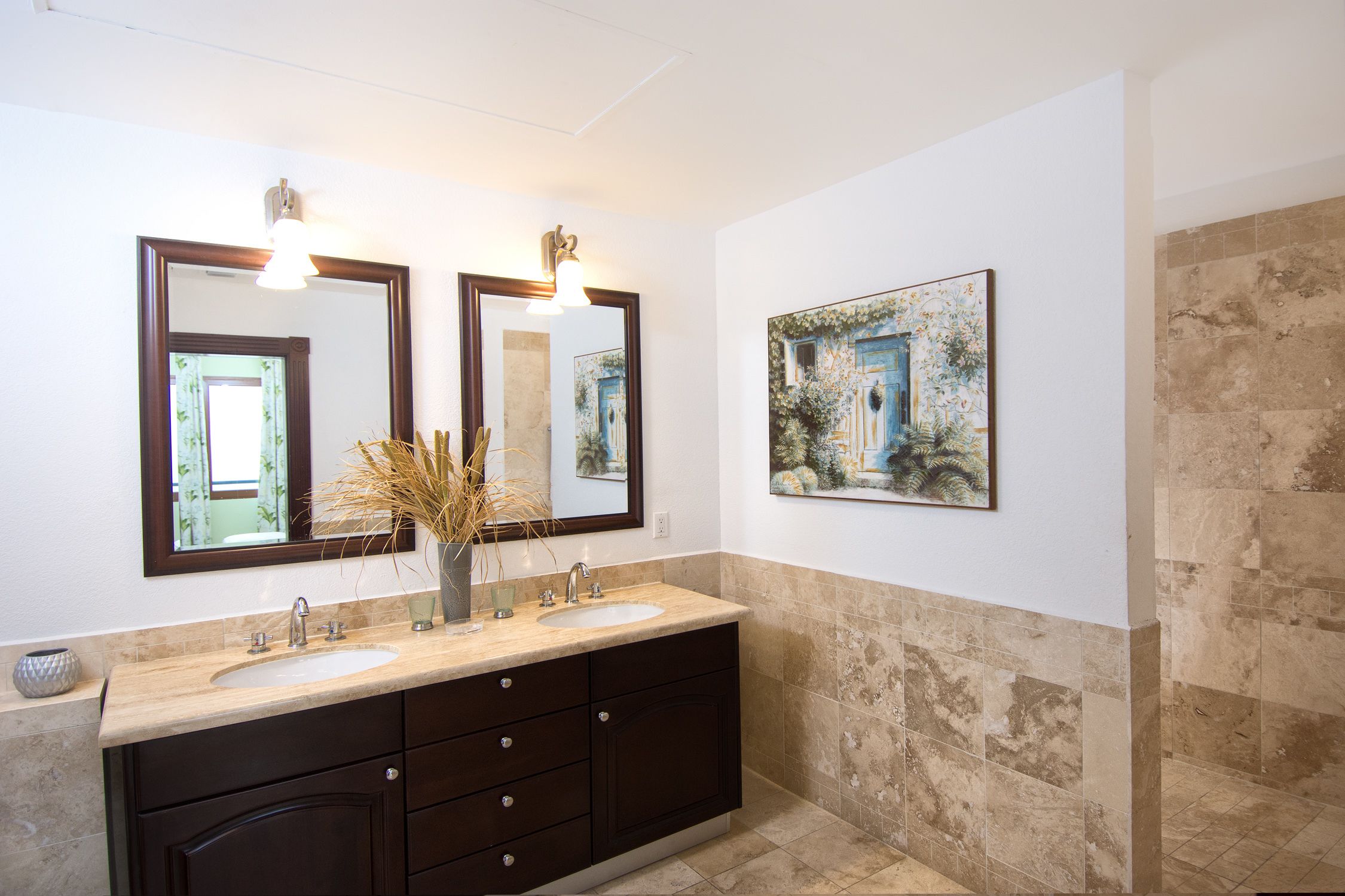 A bathroom with a double sink vanity, two wall-mounted mirrors, a vase with dried plants, and a framed painting on the wall.