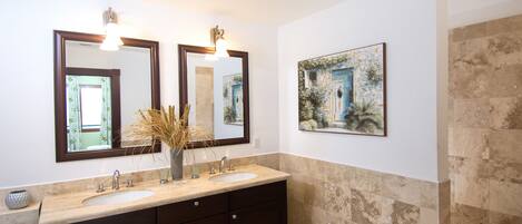 A bathroom with a double sink vanity, two wall-mounted mirrors, a vase with dried plants, and a framed painting on the wall.