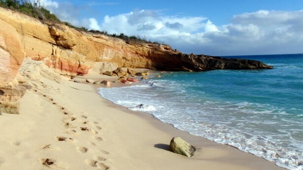 A sandy beach with footprints leading to the water, a rocky cliff, and a clear blue sky. - Artimino by Island Properties Online (Lowlands)