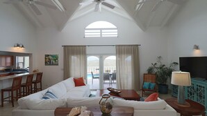 Ampla sala de estar com pé direito alto, uma grande janela e vista para o exterior. Há um sofá, uma mesa de centro e uma TV.
