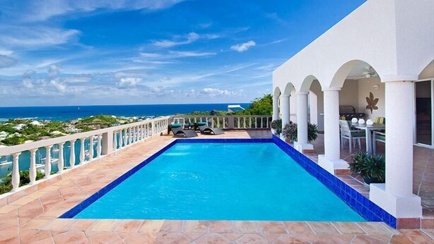 Piscina com vista para o oceano, área de varanda coberta e um edifício branco com arcos.
