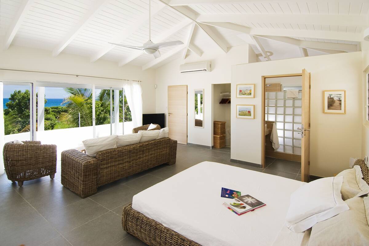A spacious bedroom with a large bed, two wicker sofas, and a view of the sea through large windows.