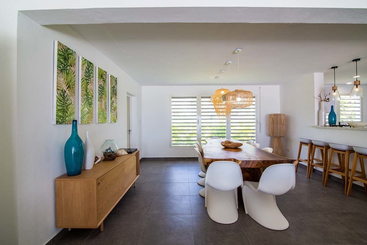 Un espace de salle à manger moderne avec une table en bois, des chaises blanches et un banc en bois. On y trouve des œuvres d’art accrochées au mur, un luminaire suspendu et une fenêtre avec des stores.